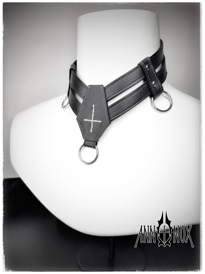 May include: A black leather choker with a cross pendant and silver metal rings. The choker has a layered design and is displayed on a white mannequin head. The brand name "ANN INOX" is visible at the bottom.