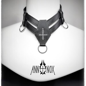 May include: Black leather choker with a coffin-shaped pendant and a cross stitched in white. The choker has two parallel straps and metal rings. The brand name "ANN NOX" is printed in black below the choker.