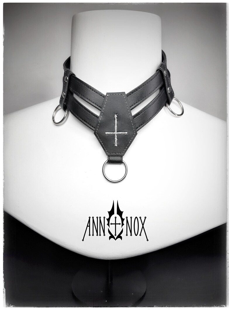 May include: Black leather choker with a coffin-shaped pendant and a cross stitched in white. The choker has two parallel straps and metal rings. The brand name "ANN NOX" is printed in black below the choker.