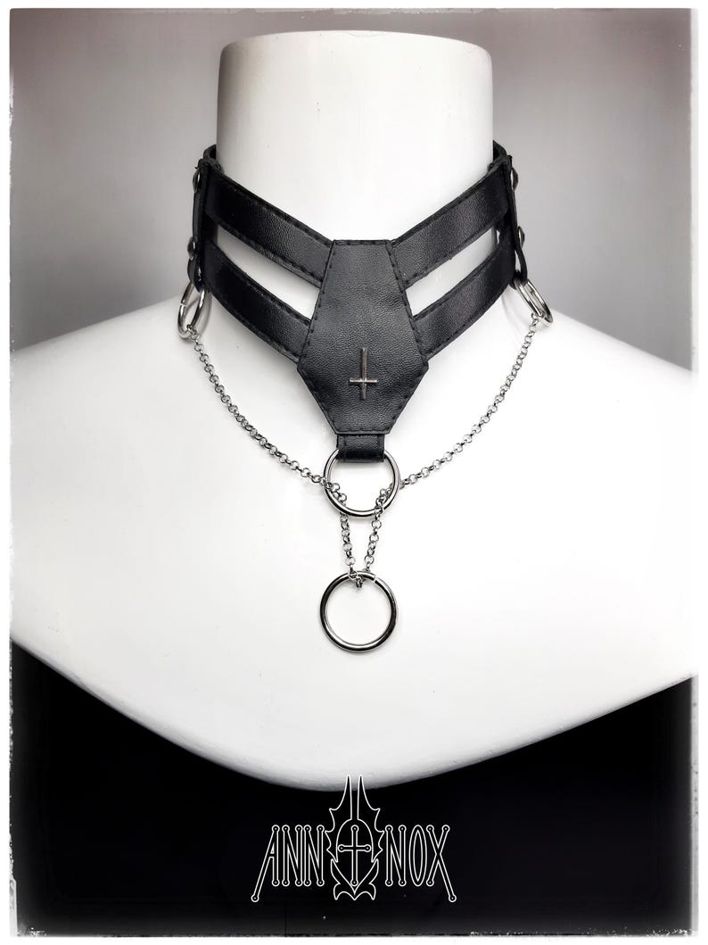 May include: Black choker necklace with a silver chain and rings. The choker features a geometric design with a cross symbol. The brand name "ANN NOX" is displayed at the bottom.