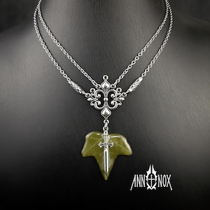 Könnte beinhalten: Silberfarbene Halskette mit Doppelketten-Design. Der Anhänger zeigt ein grünes Blatt mit einem kleinen Schwert und ein dekoratives silbernes Filigranelement. Der Markenname "ANN NOX" ist unten sichtbar.
