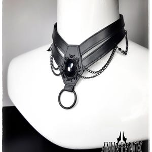 May include: Black choker necklace with a central oval-shaped black gem and decorative metalwork. The choker features a layered design with dangling chains and a large ring at the bottom. The Ann Nox logo is visible at the bottom right.
