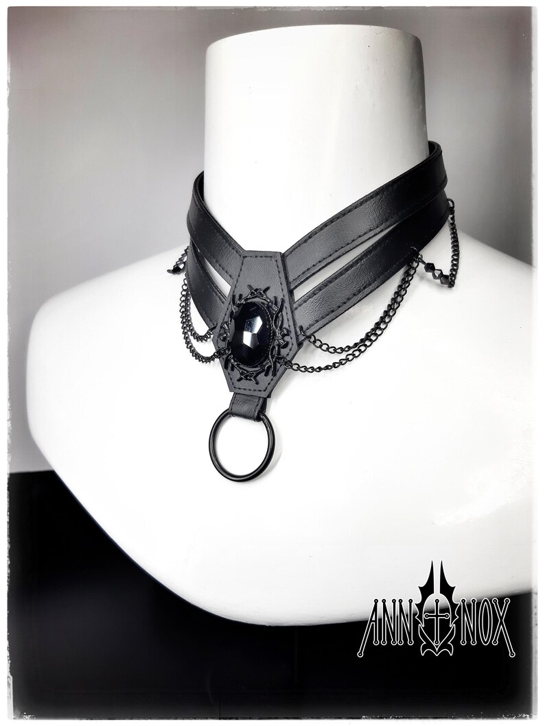 May include: Black choker necklace with a central oval-shaped black gem and decorative metalwork. The choker features a layered design with dangling chains and a large ring at the bottom. The Ann Nox logo is visible at the bottom right.
