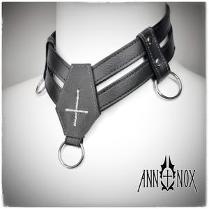 May include: Black leather choker with a coffin-shaped pendant featuring a white embroidered cross. The choker has three straps and silver metal rings. The Ann Nox logo is visible in the bottom right corner.