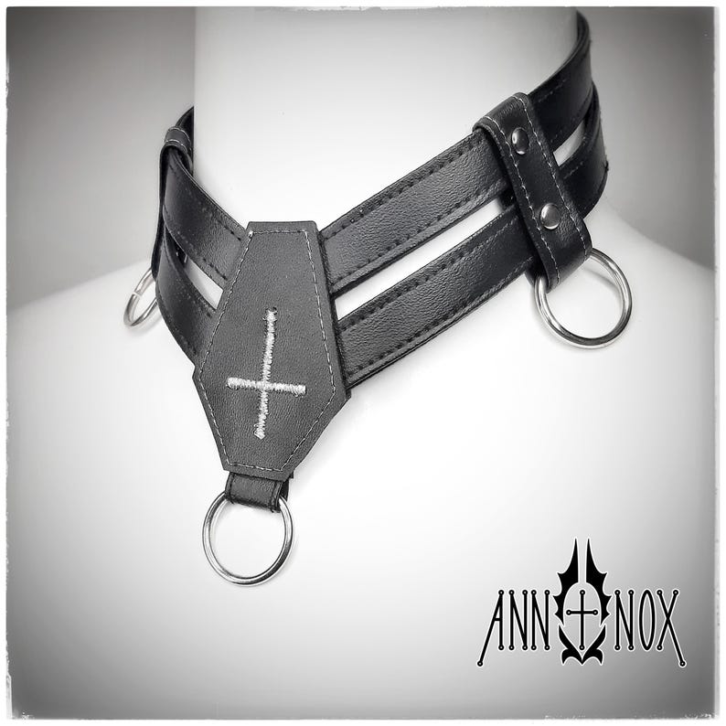 May include: Black leather choker with a coffin-shaped pendant featuring a white embroidered cross. The choker has three straps and silver metal rings. The Ann Nox logo is visible in the bottom right corner.