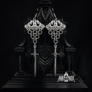 May include: A pair of silver-colored earrings with a gothic design. Each earring features an ornate top, chain links, and a sword pendant. The earrings are displayed against a dark background.