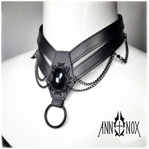 May include: Black choker necklace with a central coffin-shaped pendant featuring a large, faceted black gemstone. The pendant is adorned with intricate metalwork and dangling chains. A large black ring hangs below the pendant. The brand name "ANN NOX" is printed in black.