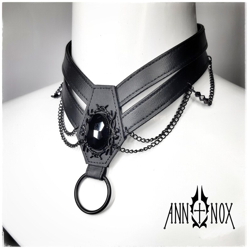 May include: Black choker necklace with a central coffin-shaped pendant featuring a large, faceted black gemstone. The pendant is adorned with intricate metalwork and dangling chains. A large black ring hangs below the pendant. The brand name "ANN NOX" is printed in black.