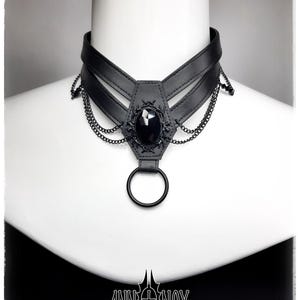 May include: Black choker necklace with a central black gem and decorative chain accents. The necklace features a leather-like band and a large ring at the bottom. The brand name "ANN NOX" is displayed in white at the bottom.