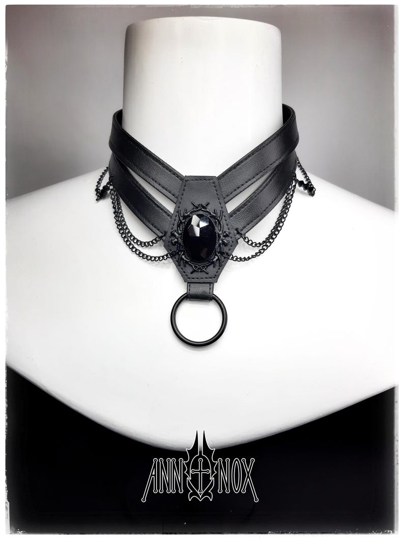 May include: Black choker necklace with a central black gem and decorative chain accents. The necklace features a leather-like band and a large ring at the bottom. The brand name "ANN NOX" is displayed in white at the bottom.