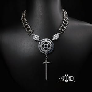 May include: A silver-toned necklace with a chain-link design and a central pendant. The pendant features a circular, openwork design and a dangling sword charm. The necklace is displayed on a black mannequin. The brand name "ANN+NOX" is visible.