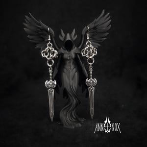 May include: Silver-toned dangle earrings with a sword and chain design. The earrings are displayed in front of a black statue of a hooded figure with wings. The Ann Nox logo is visible at the bottom right.