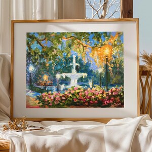May include: A framed painting of a park scene with a white fountain, lush greenery, and colorful flowers. The artwork features a warm color palette with a focus on light and shadow, creating a serene atmosphere. The artist's signature is visible in the lower right corner.