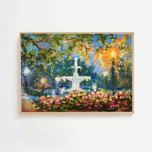 May include: An oil painting of a fountain in a park, framed in a light wood. The artwork features a white tiered fountain, surrounded by pink flowers and lush greenery. A warm light illuminates the scene, creating a serene atmosphere.