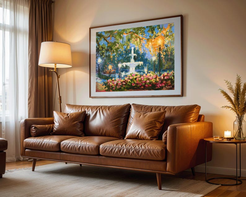 May include: A brown leather sofa with two matching throw pillows and a rolled armrest cushion. A framed painting of a fountain and flowers hangs above the sofa. A floor lamp and side table with a candle complete the living room setting.