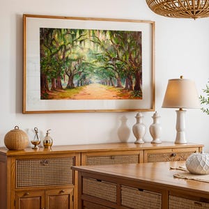 May include: A framed landscape painting of a tree-lined path with sunlight filtering through the branches. The artwork is in a wooden frame and hangs above a wooden cabinet with decorative items, including a lamp and vases. The scene evokes a sense of tranquility.