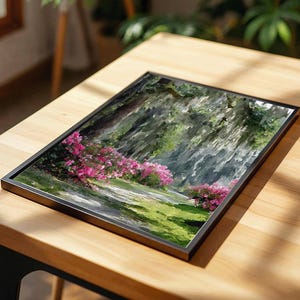 May include: A framed landscape art print featuring a pathway through a garden with pink flowering bushes and green grass. The artwork is set in a black frame and rests on a wooden table.