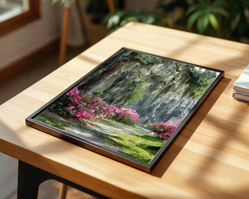 May include: A framed landscape art print featuring a pathway through a garden with pink flowering bushes and green grass. The artwork is set in a black frame and rests on a wooden table.