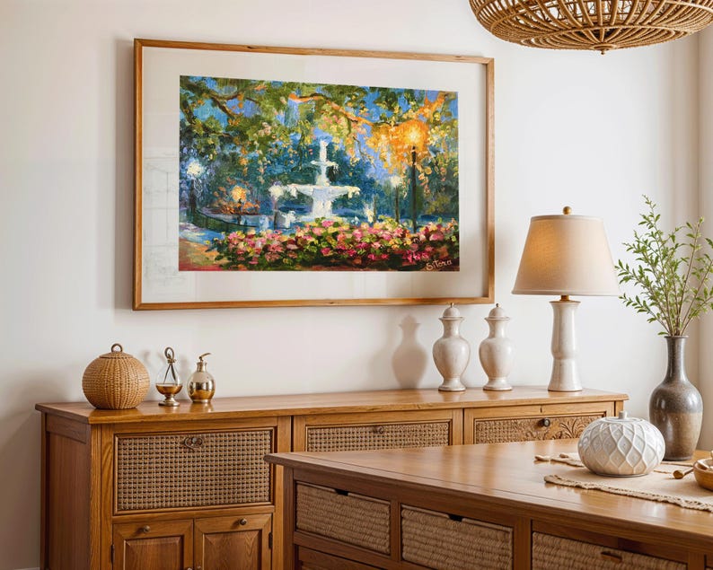 May include: A framed painting of a fountain in a garden, with pink and red flowers in the foreground. The painting is displayed above a wooden cabinet with decorative items, including a lamp and vases. A woven light fixture hangs from the ceiling.