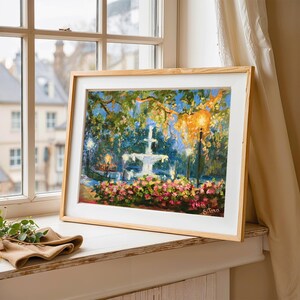 May include: A framed painting of a fountain in a park, with pink flowers in the foreground and trees overhead. The artwork is in a light wood frame, resting on a windowsill. The scene is illuminated by streetlights.