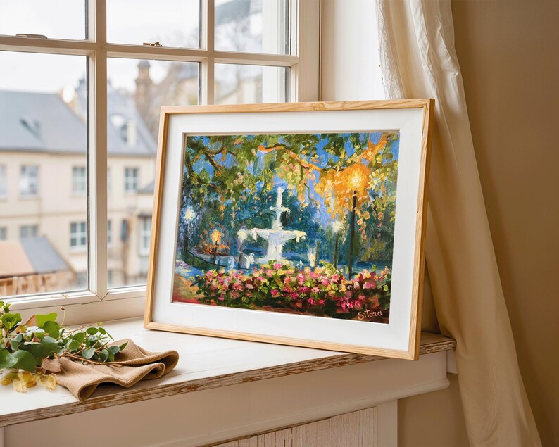 May include: A framed painting of a fountain in a park, with pink flowers in the foreground and trees overhead. The artwork is in a light wood frame, resting on a windowsill. The scene is illuminated by streetlights.
