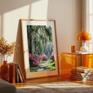 May include: A framed landscape print depicts a path through a lush forest with pink flowers. The artwork is displayed in a room with an orange side table, a lamp, and a vase with dried plants. The frame is made of wood.