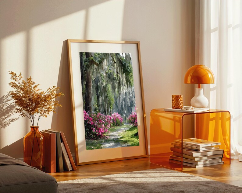 May include: A framed landscape print depicts a path through a lush forest with pink flowers. The artwork is displayed in a room with an orange side table, a lamp, and a vase with dried plants. The frame is made of wood.