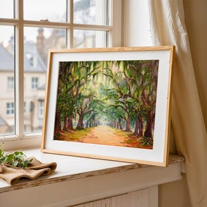 May include: A framed landscape painting depicting a tree-lined avenue. The artwork shows a path through a forest of trees with green foliage and hanging moss. The frame is light-coloured wood, and the painting is displayed on a windowsill.