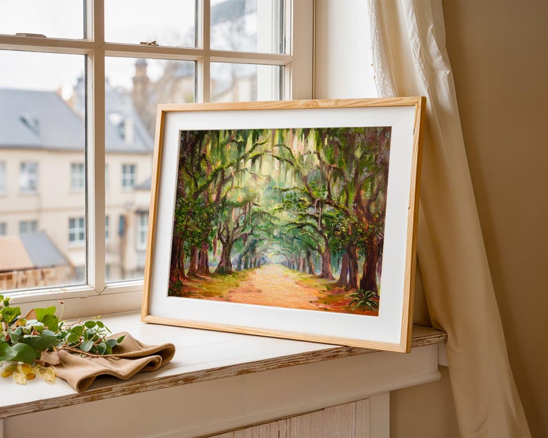 May include: A framed landscape painting depicting a tree-lined avenue. The artwork shows a path through a forest of trees with green foliage and hanging moss. The frame is light-coloured wood, and the painting is displayed on a windowsill.