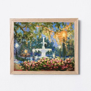 May include: An oil painting in a light wood frame depicts a fountain in a park at dusk. The fountain is white, surrounded by pink and red flowers. Trees and streetlights are visible, with a blue and yellow sky.