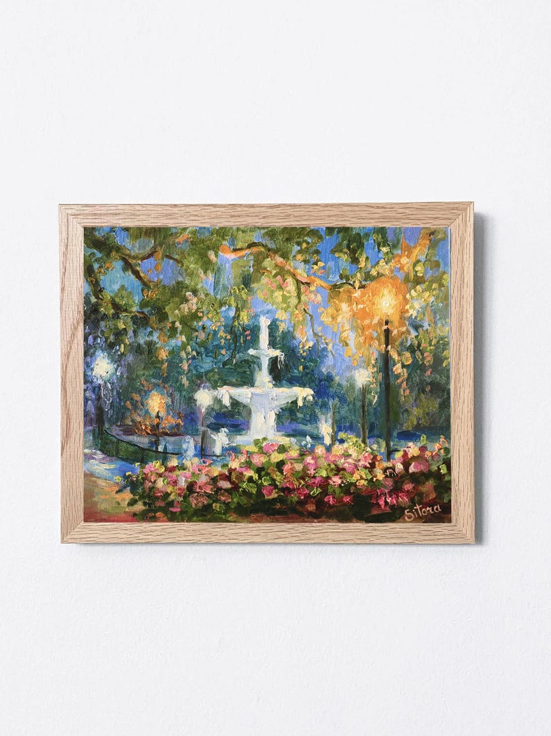 May include: An oil painting in a light wood frame depicts a fountain in a park at dusk. The fountain is white, surrounded by pink and red flowers. Trees and streetlights are visible, with a blue and yellow sky.