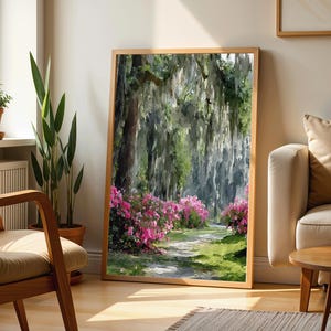 May include: A framed landscape art print depicts a path winding through a lush forest with hanging moss and vibrant pink azaleas. The artwork is in a natural wood frame, leaning against a wall near a window and a sofa.