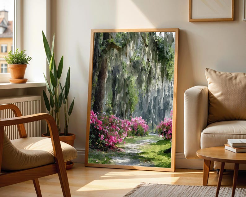 May include: A framed landscape art print depicts a path winding through a lush forest with hanging moss and vibrant pink azaleas. The artwork is in a natural wood frame, leaning against a wall near a window and a sofa.