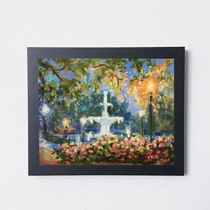 May include: A framed painting depicts a fountain in a park, surrounded by lush greenery and pink flowers. The artwork, with a dark frame, uses a palette of blues, greens, and yellows to capture the scene's ambiance. The painting is signed in the lower right corner.
