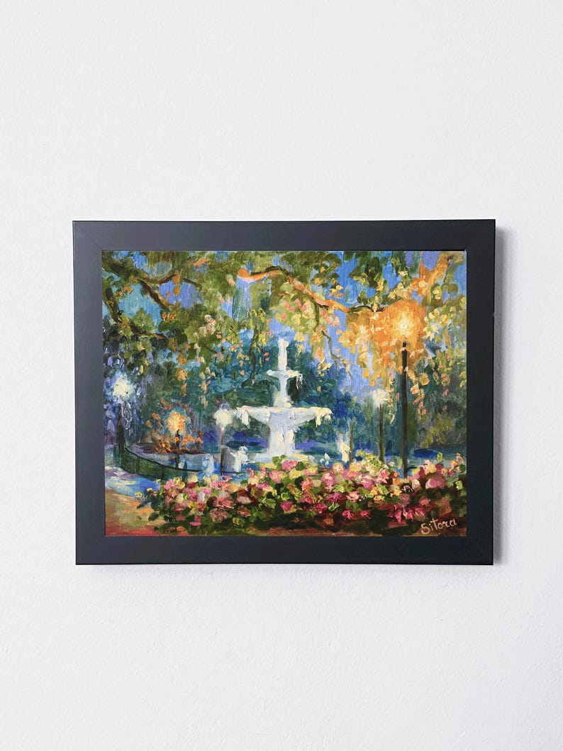 May include: A framed painting depicts a fountain in a park, surrounded by lush greenery and pink flowers. The artwork, with a dark frame, uses a palette of blues, greens, and yellows to capture the scene's ambiance. The painting is signed in the lower right corner.