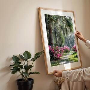 May include: A framed landscape art print featuring a pathway through a lush forest with vibrant pink flowering bushes. The artwork is in a light wood frame, adding a natural touch to the scene. The print is being held up against a neutral wall.