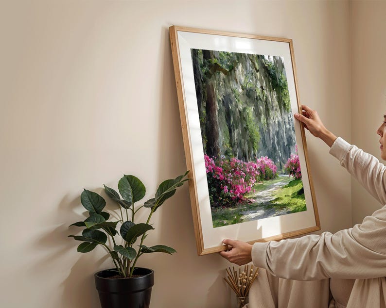 May include: A framed landscape art print featuring a pathway through a lush forest with vibrant pink flowering bushes. The artwork is in a light wood frame, adding a natural touch to the scene. The print is being held up against a neutral wall.