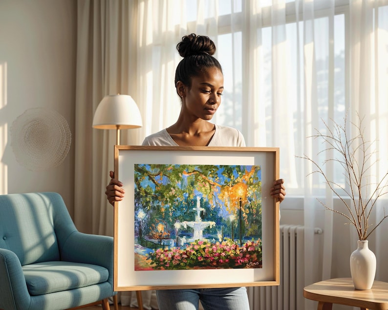May include: A framed painting of a fountain in a park, with a wooden frame. The artwork features a white fountain, lush greenery, and colorful flowers. The painting is held by a person.