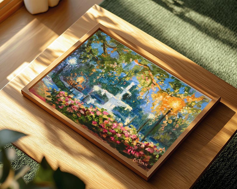 May include: A framed painting of a park scene with a white fountain, trees, and flowers. The artwork features a vibrant color palette of blues, greens, pinks, and oranges. The painting is in a wooden frame and rests on a wooden surface.