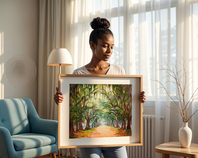 May include: A framed landscape painting showing a tree-lined path with sunlight filtering through the branches. The artwork uses a warm colour palette with greens, browns, and oranges. The frame is made of light-coloured wood.