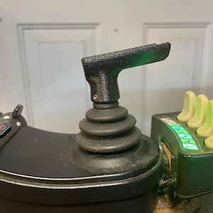 May include: Black joystick control with a textured black handle and a green light. The joystick is mounted on a black base with a series of buttons.