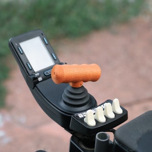 Könnte beinhalten: Ein schwarzer Joystick mit einem orangefarbenen Griff und vier weißen Knöpfen. Der Joystick ist an einem schwarzen Bedienfeld mit einem kleinen Bildschirm befestigt.