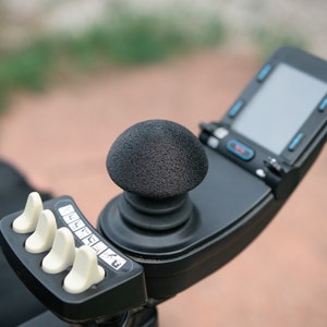 May include: Close-up of a black power wheelchair control panel. Features include a joystick with a textured black grip, a small screen, and several white buttons. The design is modern and functional.