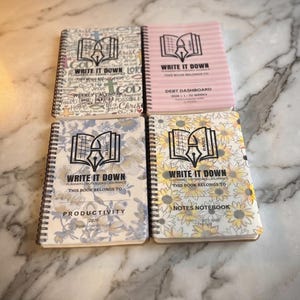 May include: Four spiral-bound notebooks with black wire binding. The top left features religious text and symbols. The top right is pink with horizontal stripes. The bottom left has a blue floral pattern. The bottom right displays a yellow sunflower print.