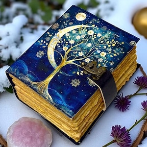 May include: A decorative journal with a blue cover depicting a gold tree, crescent moon, and stars. The pages are golden, and the journal is secured with a black leather strap and clasp. A unique gift idea.