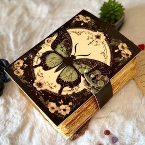 May include: A vintage-style journal with a butterfly design on the cover. The butterfly is green and black, set against a cream-colored background with floral accents. The journal has a brown leather strap and clasp closure.