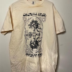 May include: A cream-colored short-sleeve t-shirt featuring a black graphic print of two skulls adorned with roses, enclosed in an oval frame. The text "GRATEFUL DEAD" appears in a stylized font above and below the central illustration on the front.