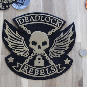 Pode incluir: Etiqueta bordada preta e dourada com uma caveira, asas, correntes e um cadeado. O texto "DEADLOCK REBELS" está bordado na etiqueta.