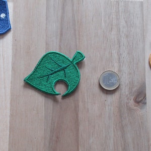 Pode incluir: Patch de folha verde bordado com um caule castanho e um furo no centro. O patch está sobre uma superfície de madeira castanho claro.