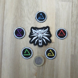 Könnte beinhalten: Sechs gestickte Patches mit Symbolen aus dem Videospiel The Witcher 3: Wild Hunt. Die Patches sind rund und zeigen einen Wolfskopf, ein Dreieck und ein Symbol, das wie ein stilisierter Buchstabe 'Z' aussieht.